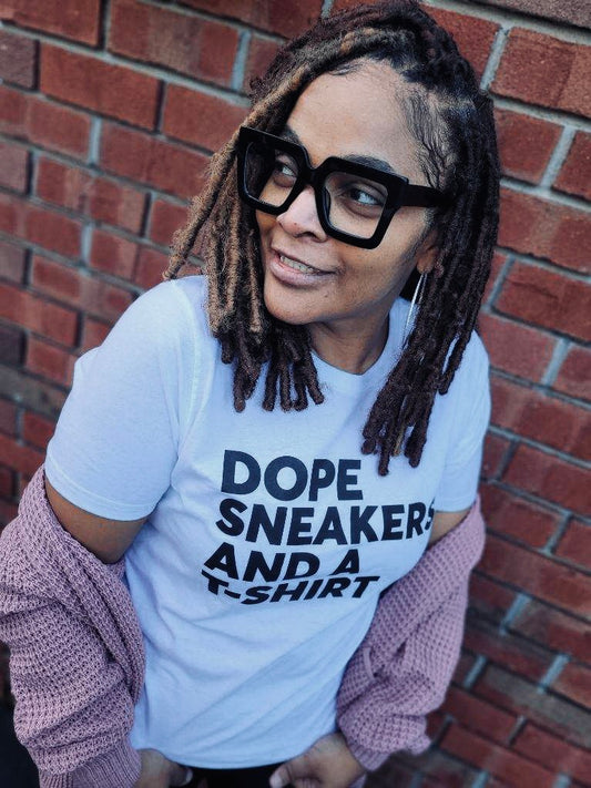 White 100% cotton tee shirt.  Model showing top part of torso .  Black lettering that says Dope Sneakers And A T-Shirt.  Model also wearing rose pink waffle cotton sweater.  